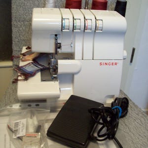 Può includere: Una macchina da cucire overlock Singer bianca con bobine di filo rosso e nero. La macchina ha un pedale e un sacchetto di plastica trasparente con aghi. Il logo Singer è visibile sul davanti.