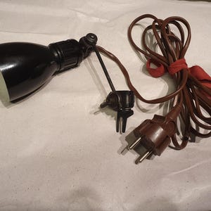 Könnte beinhalten: Ein schwarzer Klemmstrahler mit weißem Innenraum, ausgestattet mit einem verstellbaren Gelenk und einem langen braunen Stromkabel mit zweipoligem Stecker. Das Kabel ist mit einem roten Stoffband gebündelt und die Klemme hat eine schwarze Flügelmutter.
