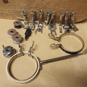 Op de afbeelding: Een verzameling zilverkleurige naaimachine-accessoires, waaronder diverse metalen naaivoetjes, drie metalen spoeltjes met garen, een spoelhuis, een zwarte schroevendraaier en twee ronde borduurringen met een lange metalen bevestigingsarm.