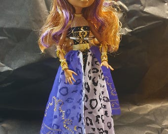 May include: A Monster High doll with tan skin, cat ears, and long, wavy auburn hair with purple streaks. She wears a black and gold top, a blue and white patterned skirt, and gold bracelets. The doll is standing on a white base.