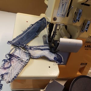 May include: A close-up of a yellow and white sewing machine with a piece of blue denim fabric and a piece of patterned fabric being sewn. The machine has a label that reads "NEEDLES" and "JL-2 BL-4".