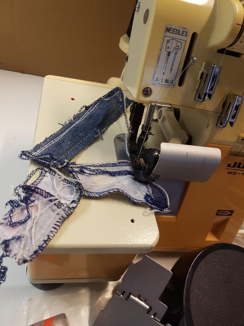 May include: A close-up of a yellow and white sewing machine with a piece of blue denim fabric and a piece of patterned fabric being sewn. The machine has a label that reads "NEEDLES" and "JL-2 BL-4".