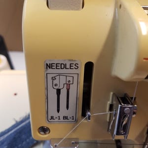 May include: Close-up of a sewing machine with two needles and a thread guide. The machine is beige and has a label that says "NEEDLES" and "JL x 1 BL x 1".