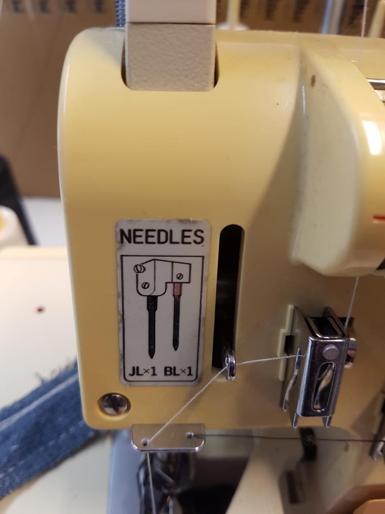 May include: Close-up of a sewing machine with two needles and a thread guide. The machine is beige and has a label that says "NEEDLES" and "JL x 1 BL x 1".