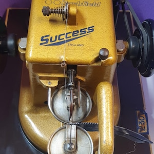 Peut inclure: Une machine à coudre industrielle vintage Allbook & Hashfield Success England de couleur dorée avec une finition texturée. La machine présente un logo bleu proéminent, un mécanisme de tension en métal, une aiguille et deux pieds presseurs circulaires.