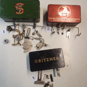 May include: A collection of vintage sewing machine parts and accessories, including metal boxes with the Singer, Kochs Adler, and Gritzner logos. Various attachments, bobbins, and presser feet are scattered around the boxes, suggesting a collection of sewing tools.