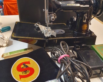 Puede incluir: Una máquina de coser Singer negra con detalles dorados, acompañada de un maletín de transporte negro a juego, un pedal negro con cable, tres bobinas de metal, una herramienta circular de metal y un parche de tela negro con una S roja.