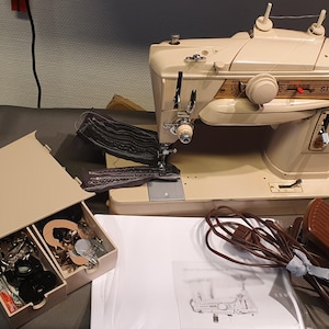 Peut inclure: Machine à coudre Singer vintage crème avec des accents dorés. L'image présente la machine, une boîte de rangement beige avec des accessoires, une pédale et un manuel. La machine a un design classique.