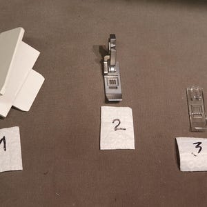 Op de afbeelding: Een assortiment naaimachineonderdelen, waaronder een naaivoet en andere hulpstukken. De afbeelding toont een wit plastic onderdeel, een metalen naaivoet en twee andere metalen hulpstukken. Elk item is genummerd op een stuk papier.