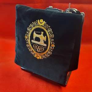May include: A black sewing machine cover with a gold embroidered design. The design features a sewing machine within an ornate oval frame. The cover is made of a soft, dark fabric and is set against a red background.