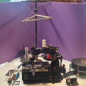 May include: A vintage black sewing machine with the text "Mader Spezial" is the focal point. The machine is set up with spools of thread, tools, and a stand. The background is a purple screen.