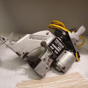 Könnte beinhalten: Eine tragbare Fischbein Modell D Sacknähmaschine mit strukturiertem silbernem Metallgehäuse, schwarzem Typenschild und einem leuchtend gelben Stromkabel. Im Vordergrund liegt ein Stück braunes Papier mit weißen genähten Fadenlinien.