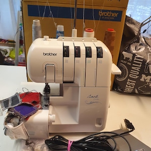 May include: A white Brother Lock 21040 serger sewing machine with a foot pedal and power cord. The machine has a clear plastic cover and a variety of stitch options. The machine is sitting on a white surface with a variety of sewing supplies.