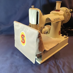 May include: A white Singer sewing machine with a grey fabric cover. The cover has a red and yellow Singer logo. The machine has a white bobbin winder and a white spool pin.