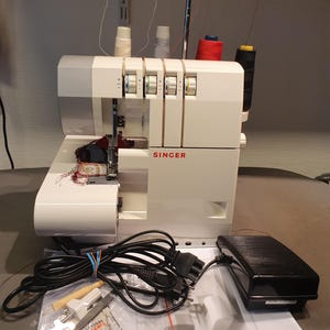 Può includere: Macchina da cucire taglia e cuci Singer bianca, con il marchio visibile. Presenta più rocchetti di filo, un pedale e accessori. La macchina è progettata per cuciture overlock, comunemente utilizzate nella produzione di abbigliamento.