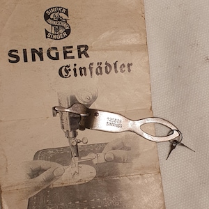May include: A metal Singer needle threader tool with the number 120328 and the word SIMANCO stamped on the handle. The tool is placed on a vintage paper instruction manual featuring the Singer logo and the text Singer Einfädler.