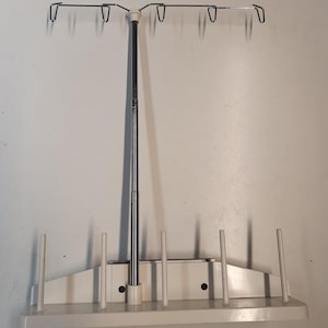 Puede incluir: Un soporte de hilo de coser blanco y plateado. El soporte tiene una barra horizontal con ganchos para sujetar los carretes de hilo. La base tiene varias clavijas verticales para sujetar los carretes. El soporte está diseñado para sujetar múltiples carretes de hilo para coser o manualidades.