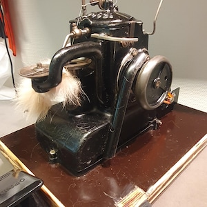 Pode incluir: Uma máquina de costura preta vintage com um grande volante e uma bobina de linha branca. A máquina está montada numa base de madeira com um pedal visível em primeiro plano. A máquina tem um braço curvo e vários componentes metálicos.