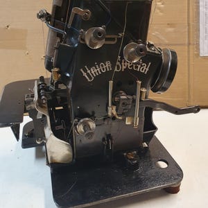 Peut inclure: Machine à coudre industrielle Union Special noire vintage. La machine a une base rectangulaire avec des pieds arrondis et une série de bobines et de leviers. Les mots "Union Special" sont imprimés sur le côté de la machine.