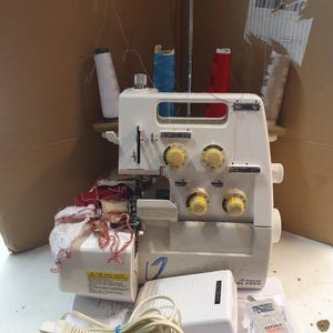 Può includere: Macchina da cucire overlock bianca a 4 fili con pedale, cavo di alimentazione e diverse bobine di filo. La macchina ha un coperchio in plastica trasparente e un'etichetta con la scritta "4-Thread Differential Feed".