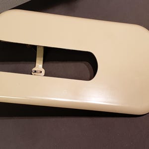 以下が含まれることがあります： クリーム色のミシンカバー。曲線的なデザインとU字型の切り欠きがあります。カバーは長方形で、角が丸く、金属製のヒンジが付いています。ヴィンテージミシン用と思われます。