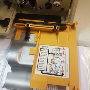 May include: A yellow sewing machine part with a diagram showing how to thread the machine. The diagram includes the text "Threading MO-103" and "Note: When passing the thread to the thread tension discs, verify that the thread has positively entered between the tension discs."