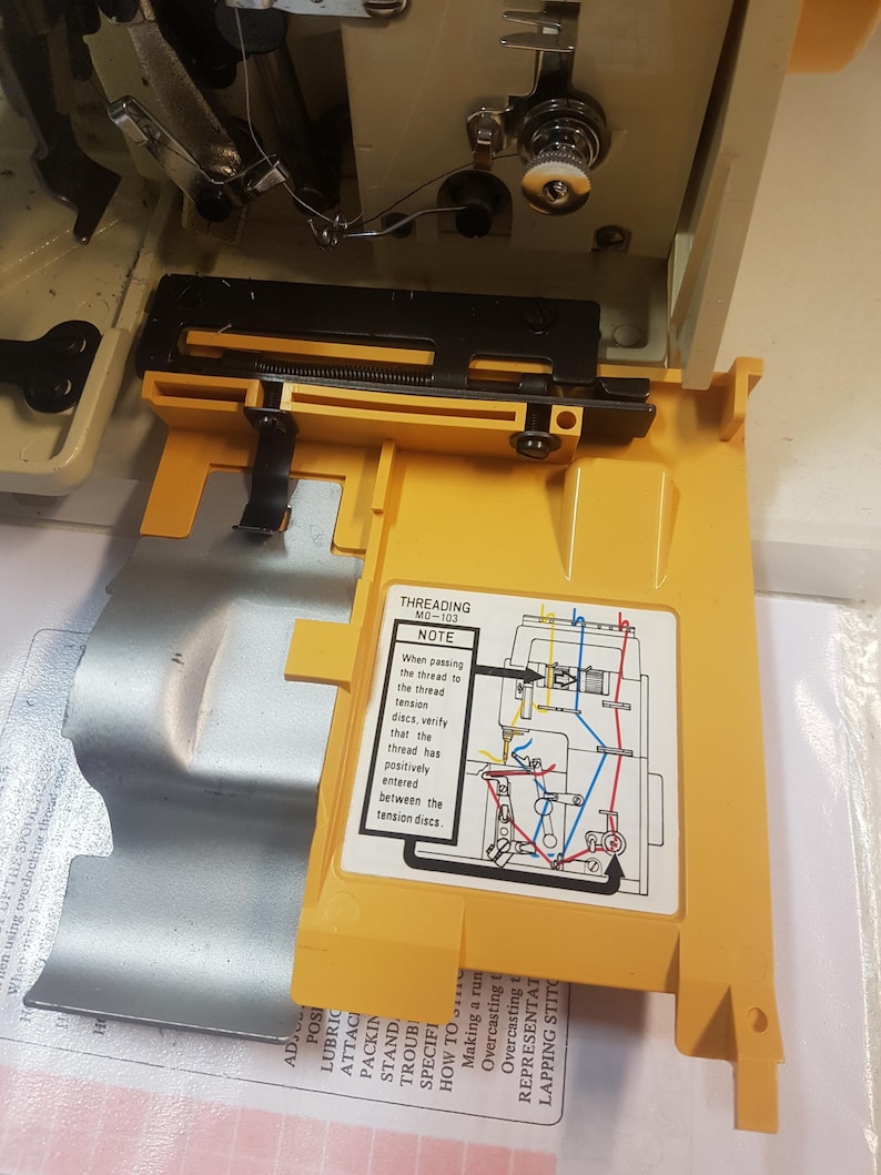 May include: A yellow sewing machine part with a diagram showing how to thread the machine. The diagram includes the text "Threading MO-103" and "Note: When passing the thread to the thread tension discs, verify that the thread has positively entered between the tension discs."