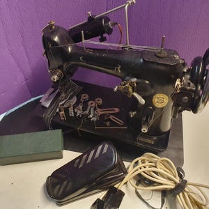May include: A black vintage Singer sewing machine with gold lettering, featuring a metal handwheel, a thread spool pin, and a needle bar. Included are several metal presser feet, a green rectangular storage box, and a black foot pedal with a cord.