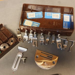 Può includere: Una collezione di parti di macchine da cucire, tra cui vari piedini, bobine e aghi. Le parti metalliche sono disposte su una superficie grigia. Gli aghi sono in scatole blu e bianche, e le bobine in una custodia marrone.