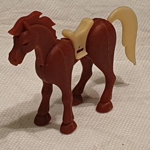 Pode incluir: Uma pequena figura de cavalo em plástico castanho com uma sela de cor creme e uma cauda a condizer na mesma cor. O cavalo está representado numa posição de pé com a cabeça ligeiramente virada para a esquerda, exibindo detalhes moldados.