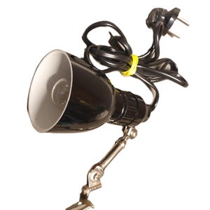 Peut inclure: Une lampe de bureau noire avec un abat-jour intérieur blanc, un bras réglable et une prise à deux broches. La lampe est reliée à un cordon noir, fixé avec un élastique jaune. La base de la lampe a un bras articulé argenté.