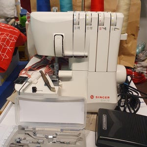 Könnte beinhalten: Eine weiße Singer S14-78 Overlock-Nähmaschine mit Fußpedal und Zubehör. Die Maschine hat mehrere Garnrollen in Blau, Rot und Creme. Das Zubehör befindet sich in einer transparenten Kunststoffbox.