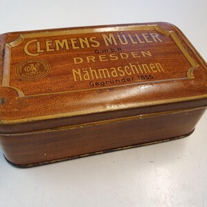 Könnte beinhalten: Eine antike Holzkiste mit goldenen Verzierungen und dem Text "Clemens Müller G.m.b.H. Dresden Nähmaschinen Gegründet 1855".