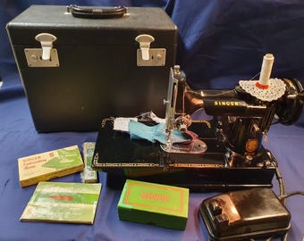 May include: A black vintage Singer sewing machine with a matching black carrying case, a foot pedal, and several original accessories including a Singer embroidery hoop box, a green Singer box, and a manual, all arranged on a blue surface.