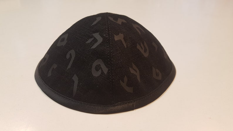 Pu&ograve; includere: Una kippah nera con un motivo di lettere ebraiche. La kippah &egrave; realizzata in un tessuto morbido e testurizzato e ha un bordo in raso nero.