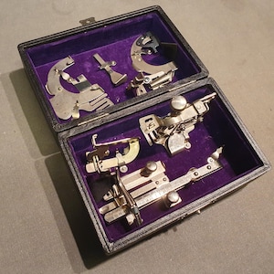 Puede incluir: Un estuche negro abierto con un interior de terciopelo púrpura contiene varios accesorios de máquina de coser de metal vintage. Los accesorios son de varias formas y tamaños, con palancas, diales y diseños intrincados.