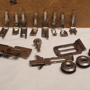 May include: A collection of silver-toned metal sewing machine accessories including eight presser feet, three circular bobbins, a knurled thumb screw, a flat metal guide plate, and a larger adjustable metal attachment with a small screw component.