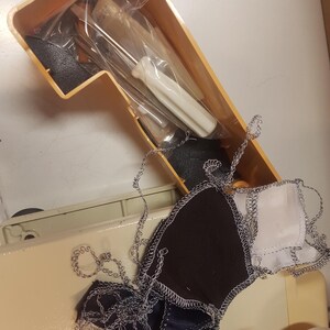 May include: A close-up of a sewing machine with a small screwdriver and a plastic bag in the accessory compartment. There are scraps of fabric with zig-zag stitching on the machine.