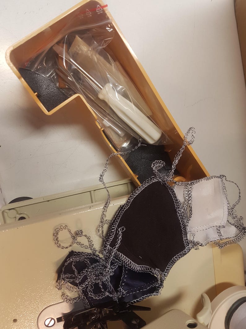 May include: A close-up of a sewing machine with a small screwdriver and a plastic bag in the accessory compartment. There are scraps of fabric with zig-zag stitching on the machine.