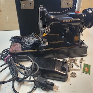 Pode incluir: Uma máquina de costura Singer preta vintage com detalhes dourados, um pedal e um cabo de alimentação. A máquina é acompanhada por vários acessórios, incluindo bobinas e um pequeno livreto de instruções. Uma mala de transporte preta está no fundo.