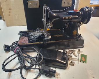 Puede incluir: Una máquina de coser Singer negra vintage con detalles dorados, un pedal y un cable de alimentación. La máquina está acompañada de varios accesorios, incluyendo bobinas y un pequeño folleto de instrucciones. Un estuche de transporte negro está en el fondo.