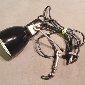Peut inclure: Une lampe de bureau noire avec une finition brillante. La lampe a un bras pivotant et un intérieur blanc. Le cordon électrique est noir et maintenu par une attache jaune. La prise est à deux broches.
