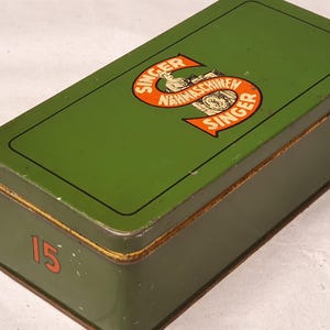Könnte beinhalten: Eine rechteckige grüne Metalldose mit einer schwarzen Umrandung auf dem Deckel. Der Deckel zeigt ein großes orangefarbenes S-förmiges Logo mit dem Text SINGER NAHMASCHINEN SINGER. Die Zahl 15 ist in Orange auf der Vorderseite der Dose aufgedruckt.