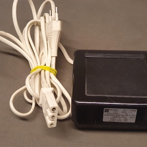 May include: A black rectangular power adapter with a white power cord and a two-prong plug. The label on the adapter reads Model 4C-125G, 220-240V, 50Hz, Matsushita Electric Ind. Co. Ltd., Made in Japan. The white cord is secured with a yellow tie.