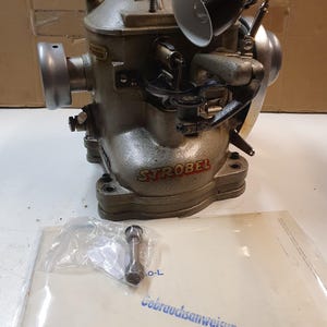 May include: A vintage silver-toned industrial machine with the brand name "STROBEL" visible. A black lamp is attached to the machine. A small part is in a plastic bag, and a manual is below the machine.