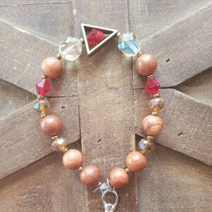 May include: A wooden beaded bracelet with a silver charm and a triangular pendant with a red gemstone. The bracelet is on a wooden background.