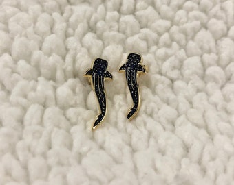 Peut inclure: Deux épingles dorées en forme de requin-baleine avec des détails en émail noir. Les épingles présentent un corps bleu foncé avec des motifs pointillés blancs et un contour doré. Les épingles sont présentées sur une surface blanche texturée.