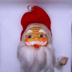 May include: A Santa Claus ornament featuring a molded face with blue eyes, rosy cheeks, and a white mustache. The figure wears a red felt hat with a gold hanging ring at the top and has a long, white, fluffy beard made of synthetic fiber material.