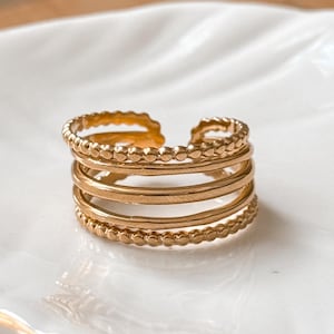 May include: A gold-colored ring with a multi-layered design. The ring features four bands, with the top and bottom bands having a beaded texture. The ring is displayed on a white, leaf-shaped dish.