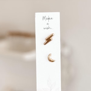May include: Gold-colored stud earrings in a lightning bolt and crescent moon design. The earrings are displayed on a white card with the handwritten text "Make a wish...". The card is held by a person.